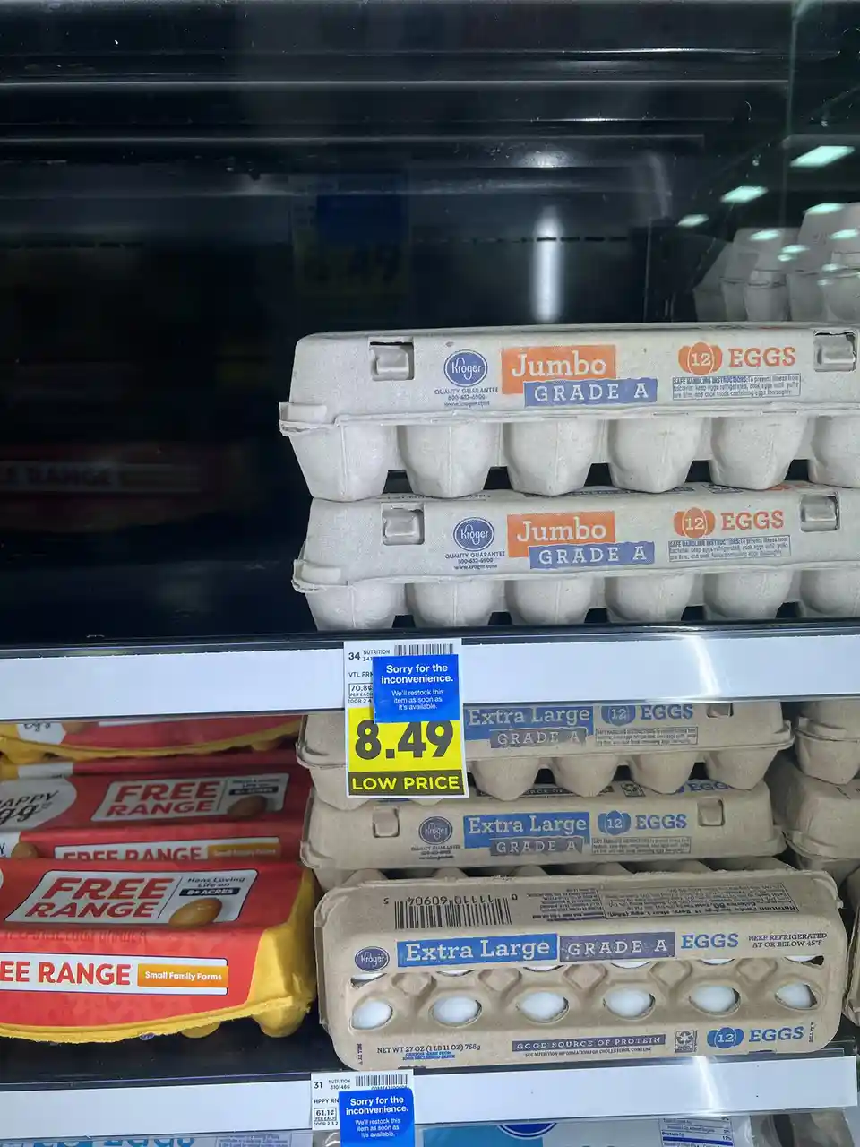 A grocery store egg shelf shows stacks of Kroger brand Jumbo and Extra Large Grade A eggs, with a prominent yellow price tag…