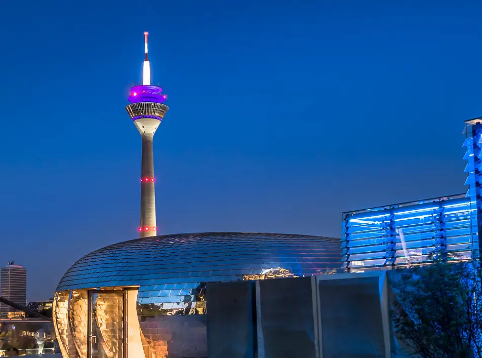 A nighttime photo of the Berlin TV Tower, illuminated with purple and red lights, standing above modern buildings with…