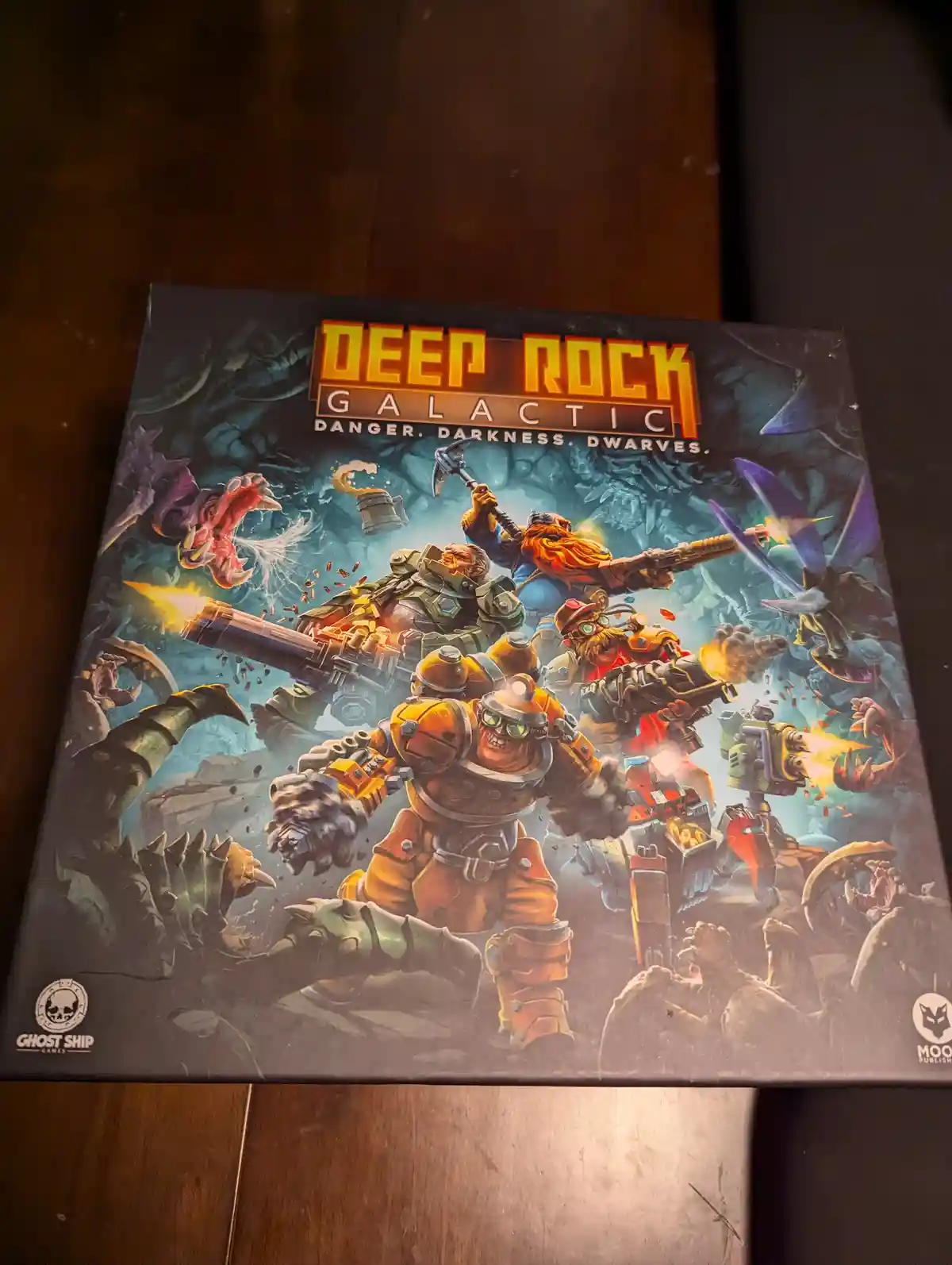 The image shows the box art for the board game 'Deep Rock Galactic', featuring four dwarf characters in heavy armor firing…