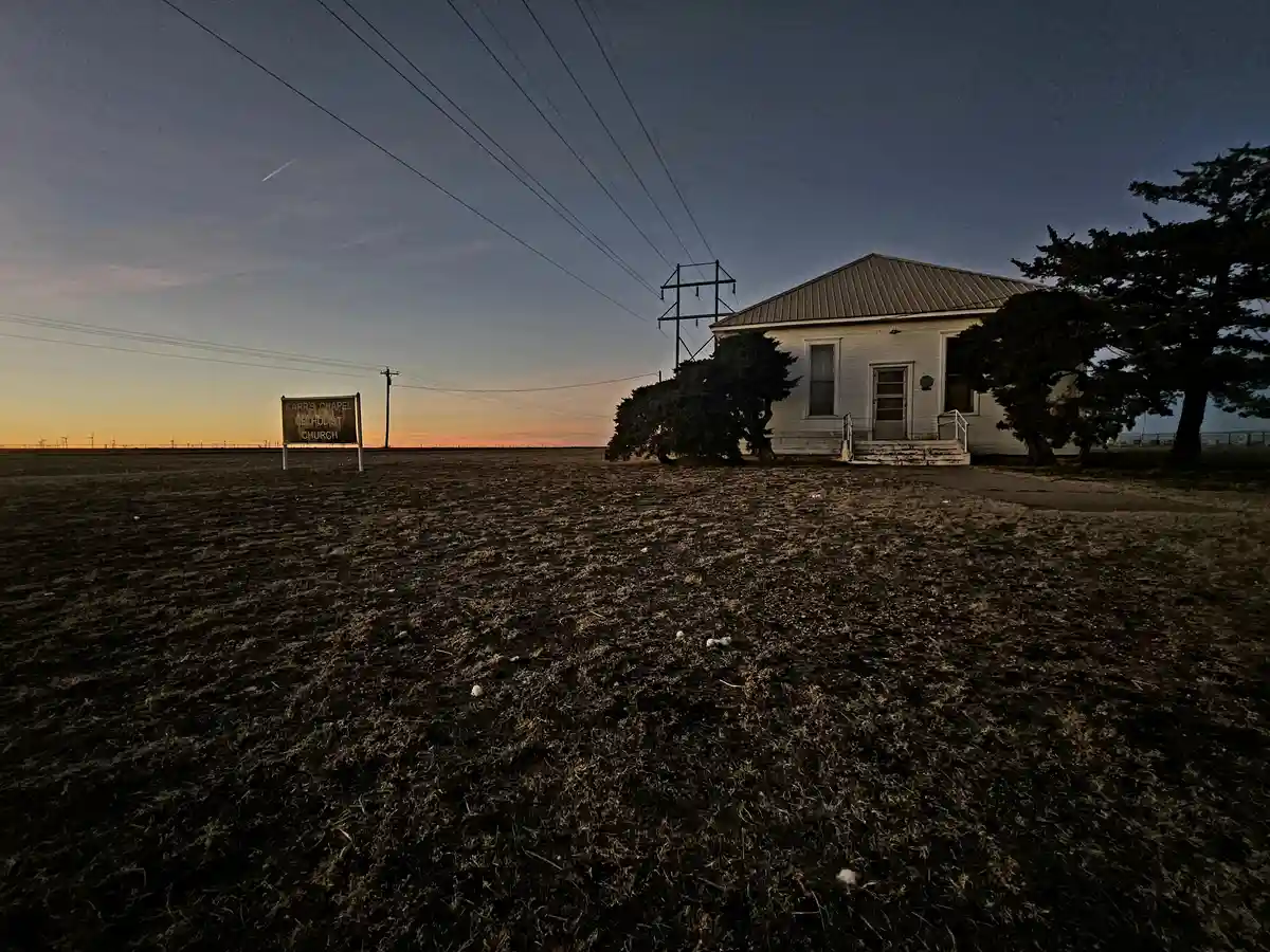 A small, white, single-story church building stands alone in a vast, flat, dry field at dusk, with a weathered sign reading…