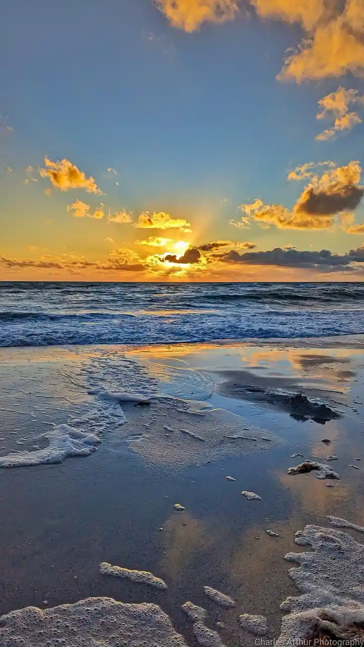 A serene beach sunset photo with golden sun rays piercing through clouds, reflecting on wet sand and gentle ocean waves. The…