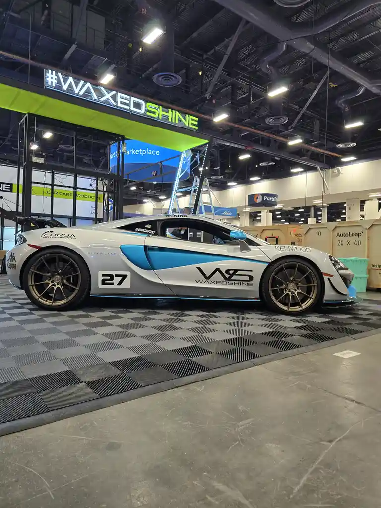 A white and blue McLaren sports car with 'WAXEDSHINE' branding and the number 27 is displayed at an auto show, parked on a…