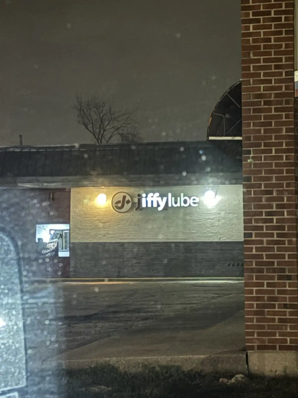 A nighttime photo taken from inside a vehicle shows a Jiffy Lube storefront with its illuminated sign visible through a…