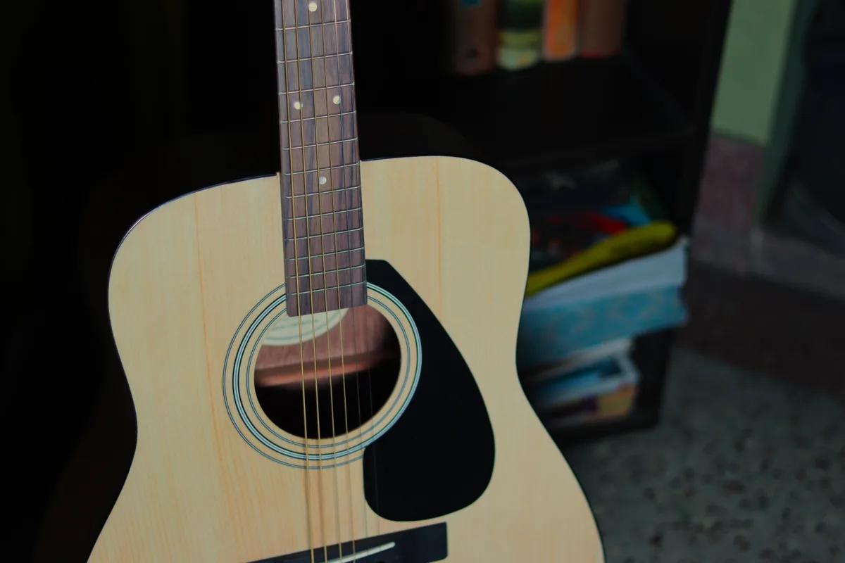 A close-up photo of a light-wood acoustic guitar with a dark fretboard and strings, positioned upright against a dark…