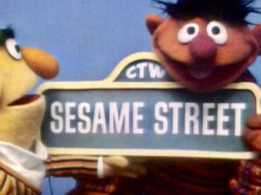 Two Sesame Street puppets, Ernie and Bert, are holding a blue street sign that reads 'SESAME STREET' with 'CTW' above it…