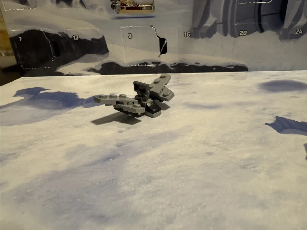 A small, gray Lego spaceship model sits on a white, textured surface resembling snow or moon dust. Behind it is a partially…