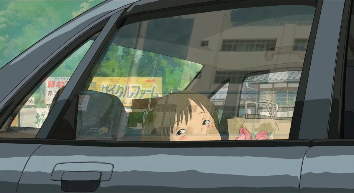 A young girl with short brown hair peeks out from the passenger side window of a car, looking up with a slightly sad or…