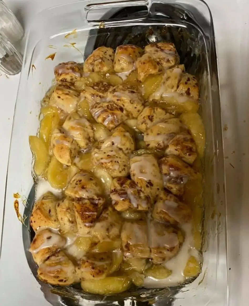 A glass baking dish contains a baked dessert with chunks of bread, sliced apples, and a white glaze, resembling apple…