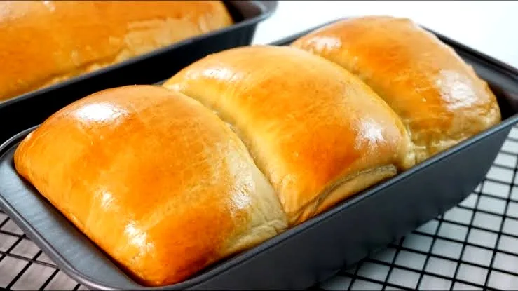 a close-up photo of three golden-brown, glossy, freshly baked loaves of bread sitting in a black loaf pan on a wire cooling…