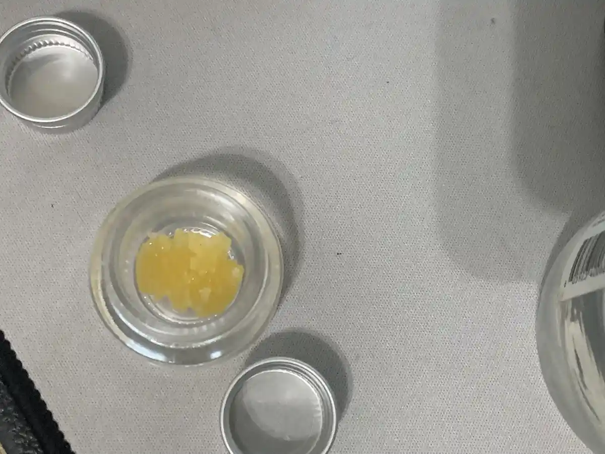 A top-down photo shows a small clear glass jar with a silver lid, open and containing a small amount of yellowish, chunky…
