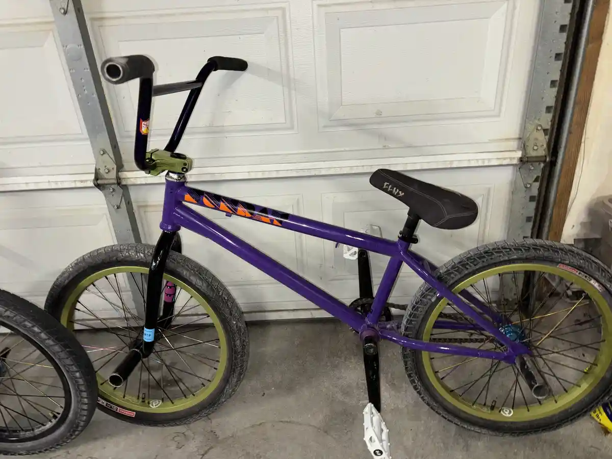 A purple BMX bike with green rims and black handlebars is parked upright in a garage in front of a white garage door. The…
