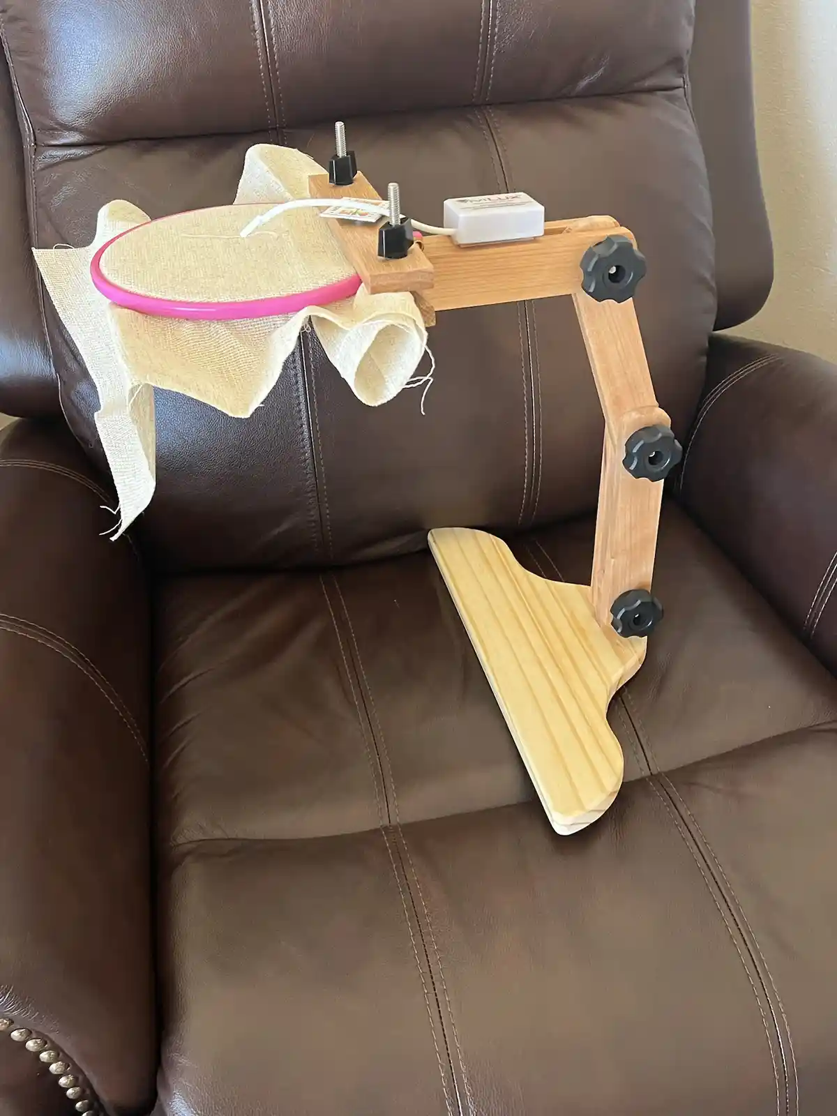 A wooden DIY stand is attached to a brown leather recliner, holding an embroidery hoop with fabric. The stand has black…