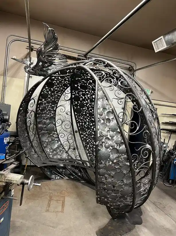 A large, ornate metal sculpture shaped like a pumpkin sits in a workshop, featuring intricate scrollwork, circular metal…