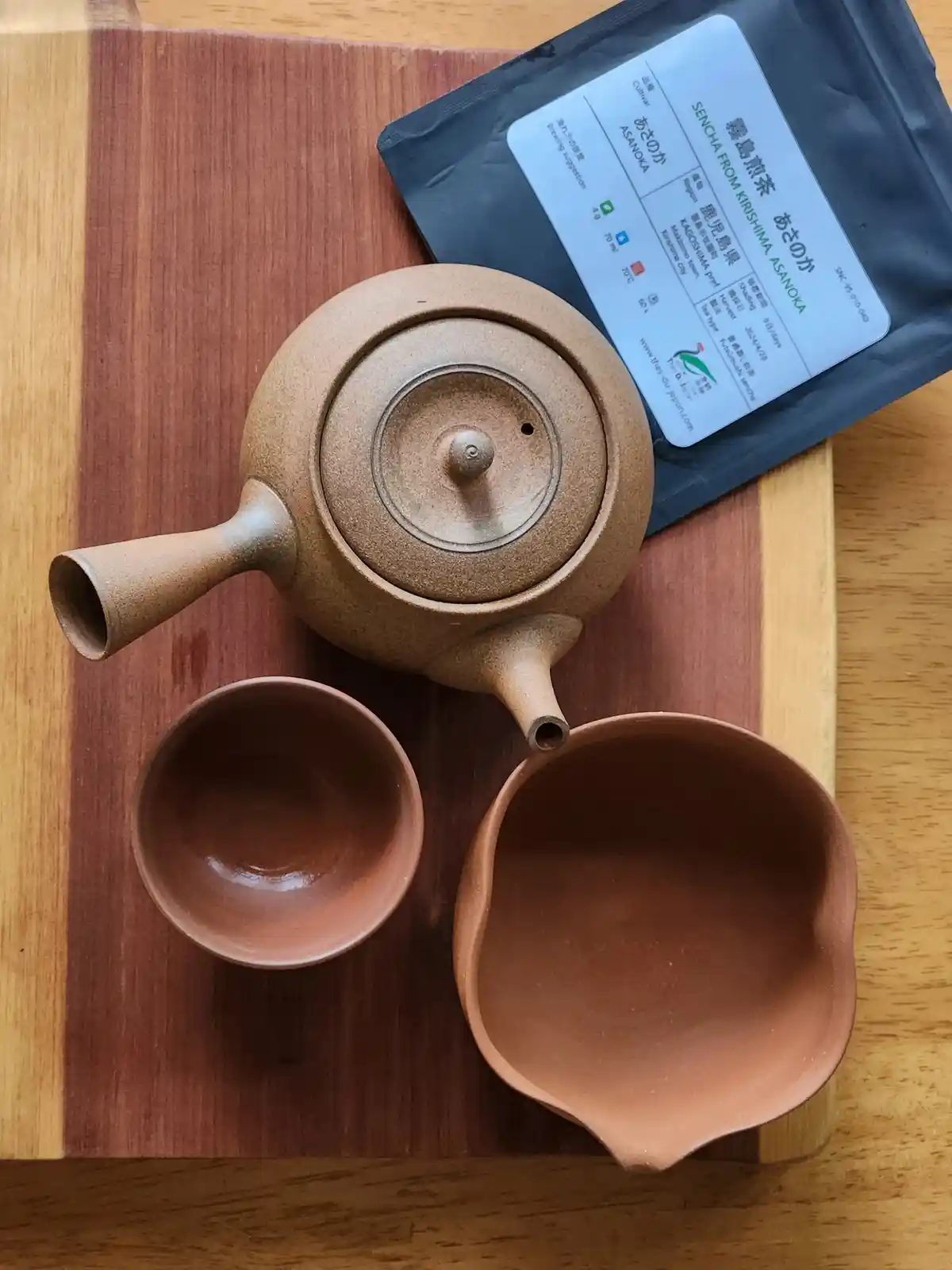 An overhead shot displays a rustic ceramic teapot, a small cup, and a larger pouring bowl arranged on a wooden board next to…