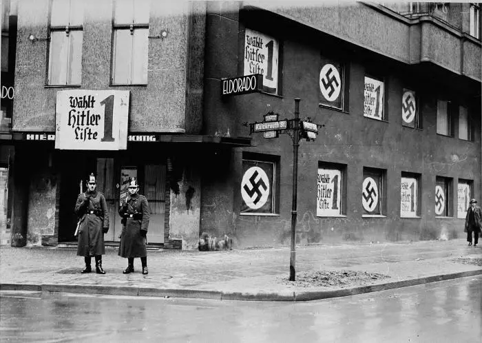 A black-and-white historical photograph shows two uniformed men standing at the corner of a street in front of a building…