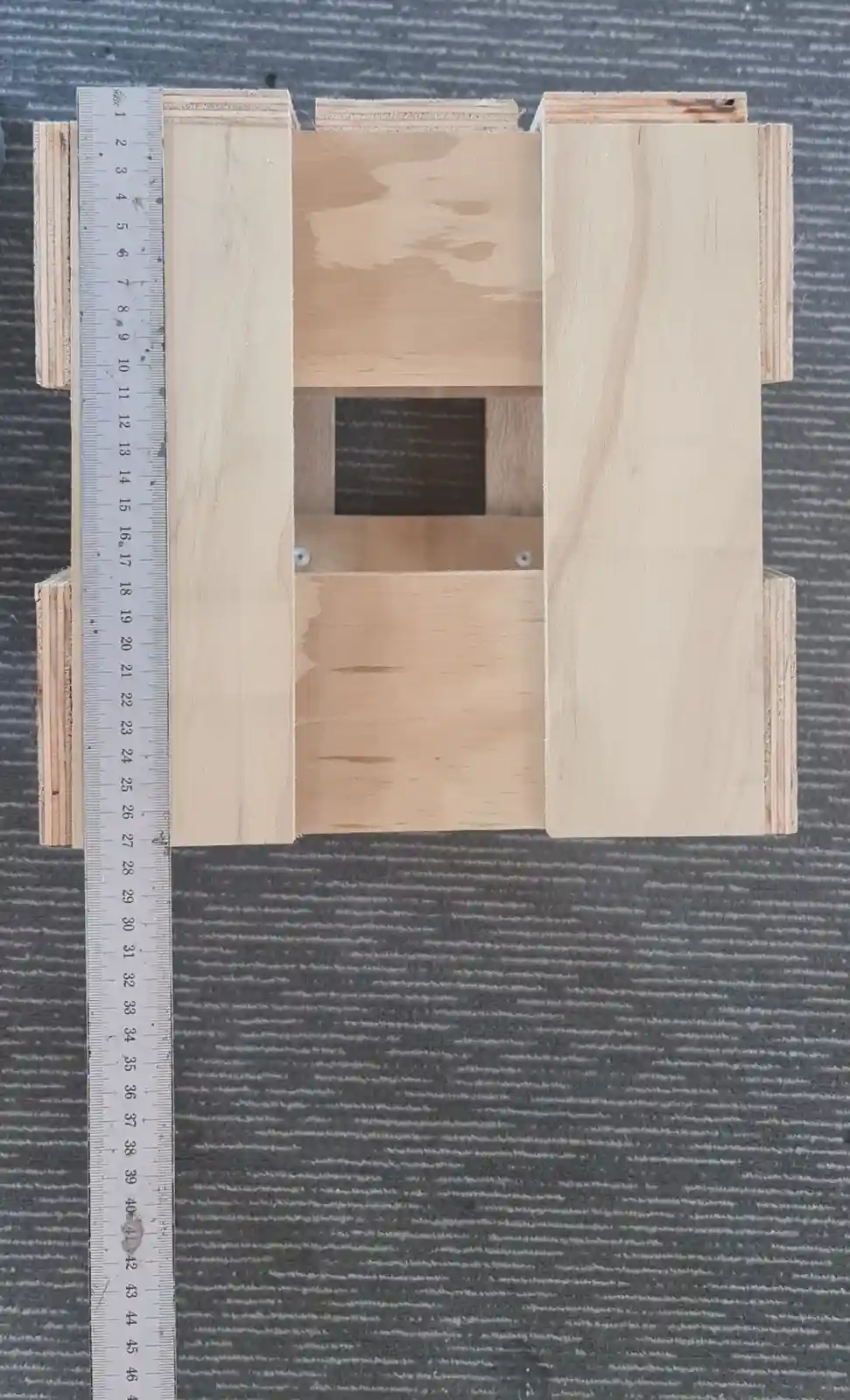 A top-down view of a wooden birdhouse frame, with a metal ruler placed vertically alongside it showing measurements up to 42…