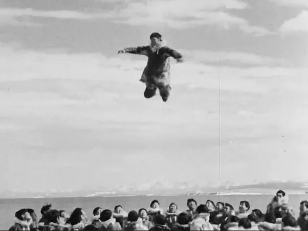 A black-and-white photo shows a man mid-air, seemingly jumping or flying above a crowd of onlookers. The crowd is seated or…