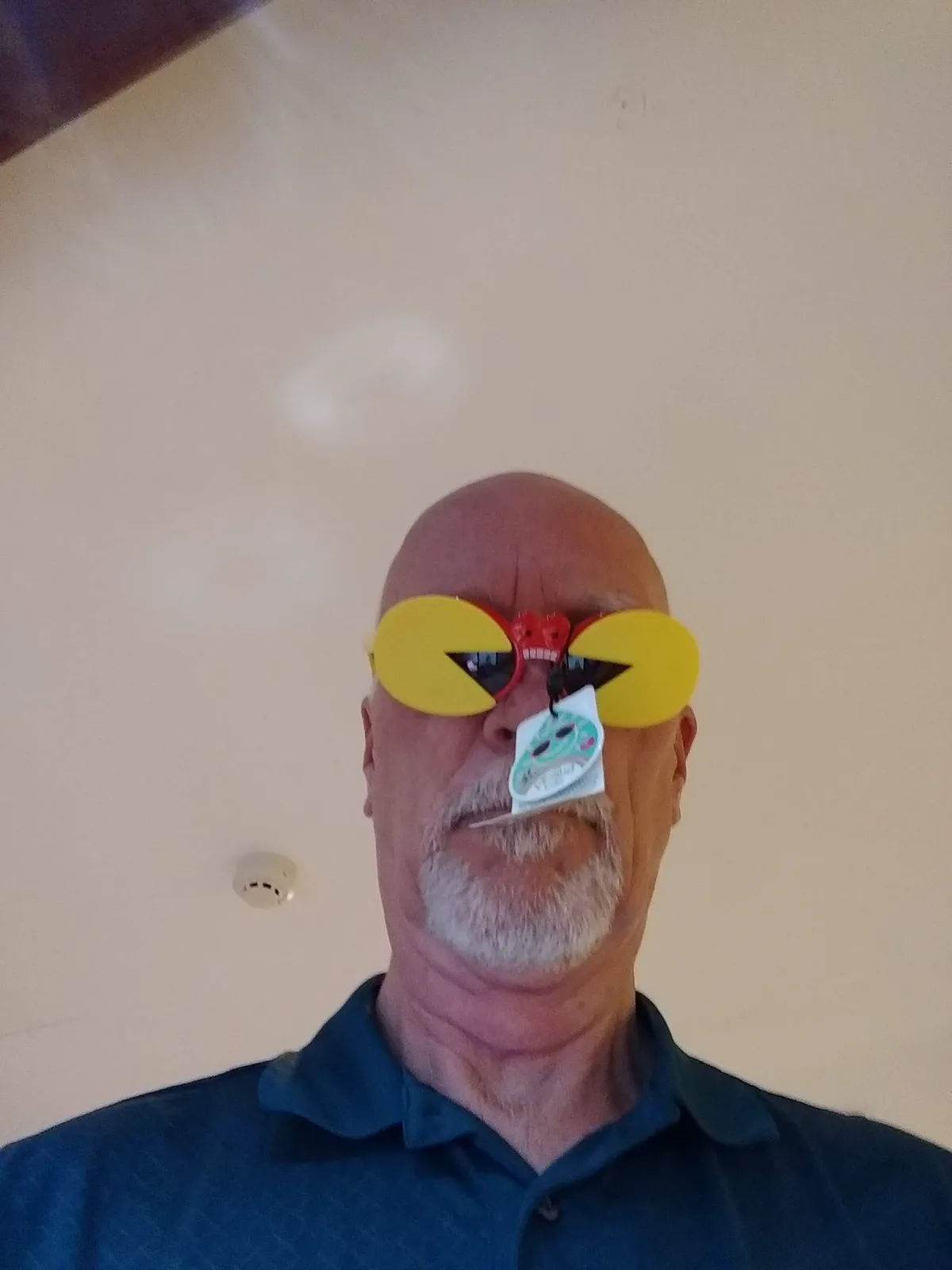 A bald man with a gray goatee wears novelty Pac-Man sunglasses with the iconic yellow mouth shape and red frame. A product…