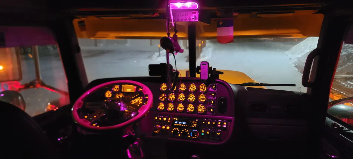 The image shows the interior of a snowplow truck at night, with a dashboard lit in purple and orange, a Texas flag hanging…
