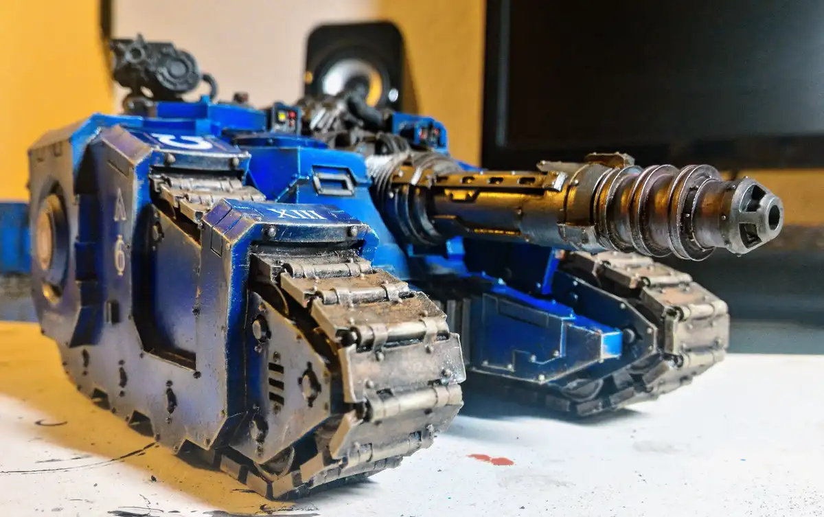 A detailed blue and silver painted model of a futuristic tank with a large, segmented cannon, sitting on a workbench with…