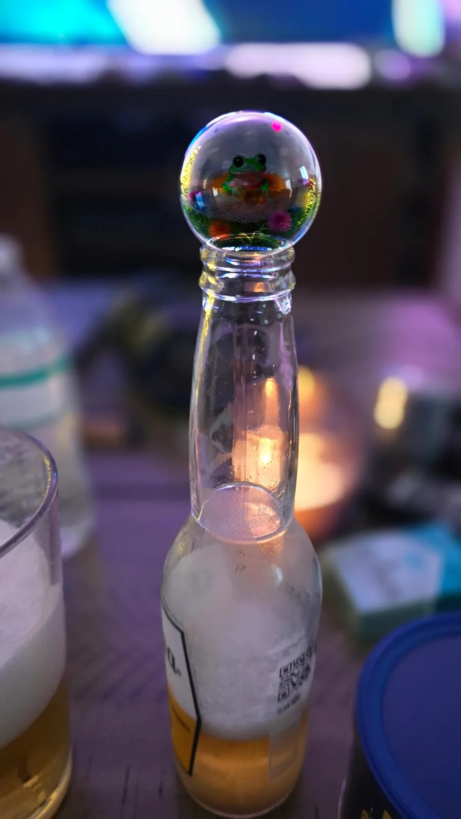 A small green frog sits inside a clear glass sphere, which is perched atop the neck of an empty beer bottle. The bottle has…