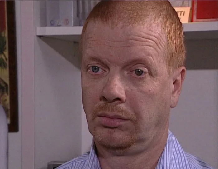 a close-up of a man with red hair and a mustache wearing a striped shirt, looking directly at the camera with a skeptical…