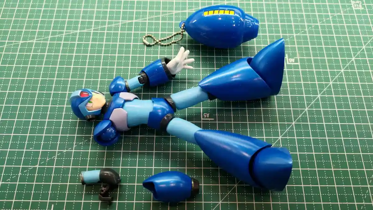 A disassembled blue Mega Man action figure lies on a green cutting mat, with its head, torso, arms, legs, and a detached…