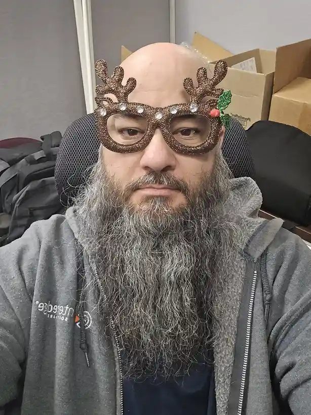 A bald man with a long grey beard sits in an office chair wearing sparkly brown reindeer antler glasses with rhinestones and…