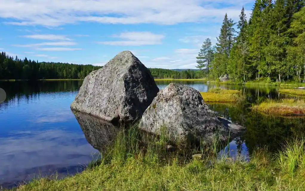 A serene landscape photo shows two large, mossy boulders sitting at the edge of a calm lake, surrounded by green grass and…