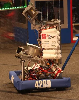 A robot with a blue padded base and the number 4269 on it, constructed from various mechanical parts, wires, and white…