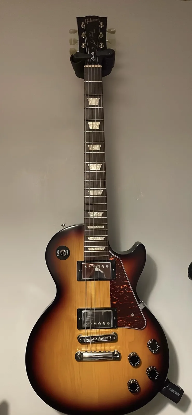 A Gibson Les Paul Studio electric guitar with a sunburst finish is mounted vertically on a plain wall. The guitar has a…