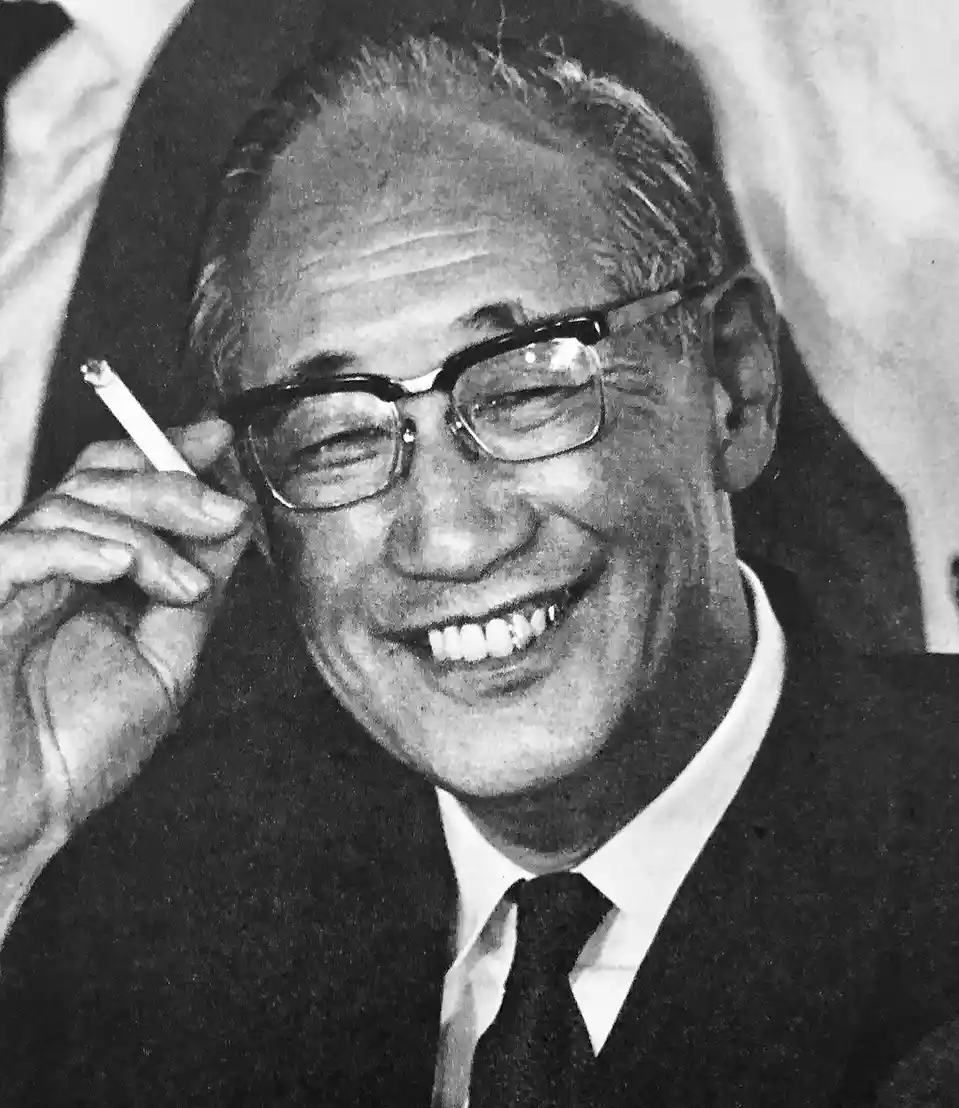 a black and white photo of an older man with glasses, wearing a suit and tie, smiling broadly while holding a cigarette near…