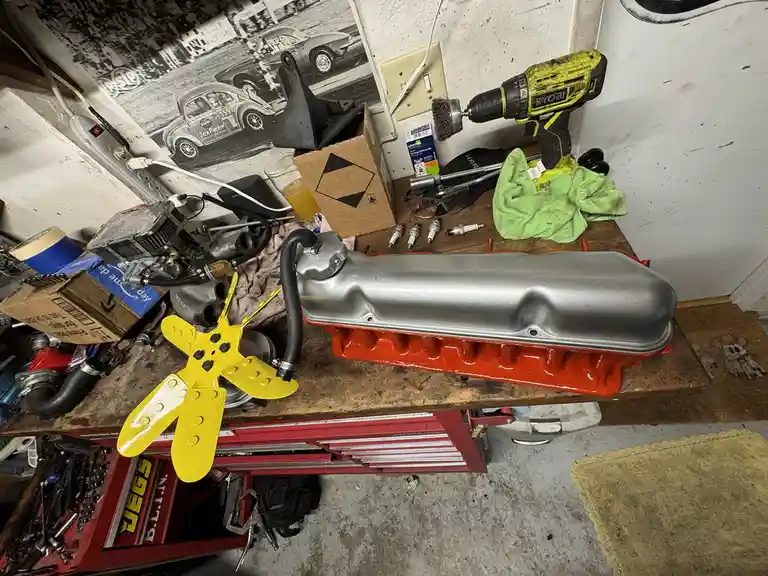 A cluttered garage workbench holds a freshly cleaned silver valve cover resting on a red engine block, surrounded by tools…