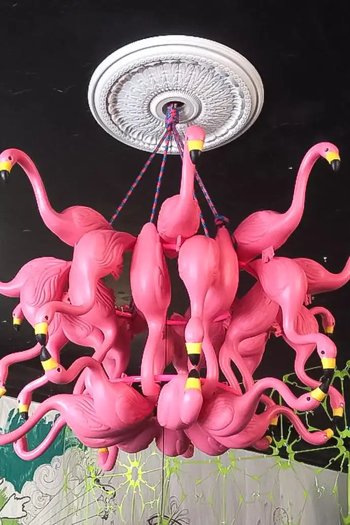 A chandelier made of numerous bright pink plastic flamingos hangs from a white ornate ceiling medallion against a dark…