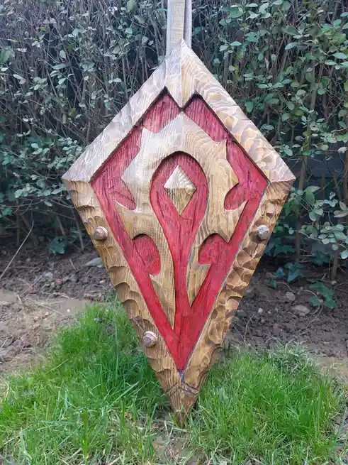 A hand-carved wooden shield with red and natural wood coloring, featuring a stylized emblem, stands upright in grass with…