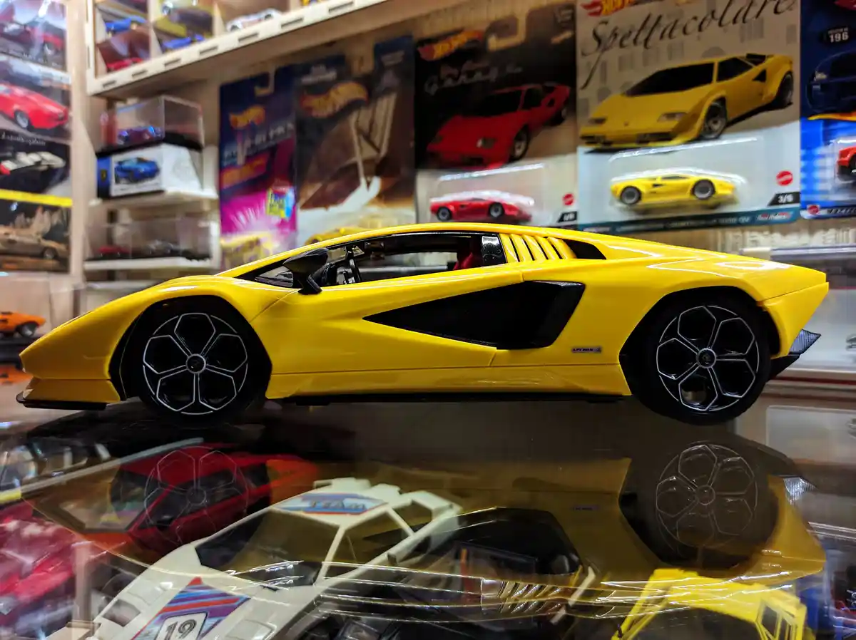 A bright yellow die-cast Lamborghini model car is displayed on a reflective surface in a toy store, surrounded by other Hot…