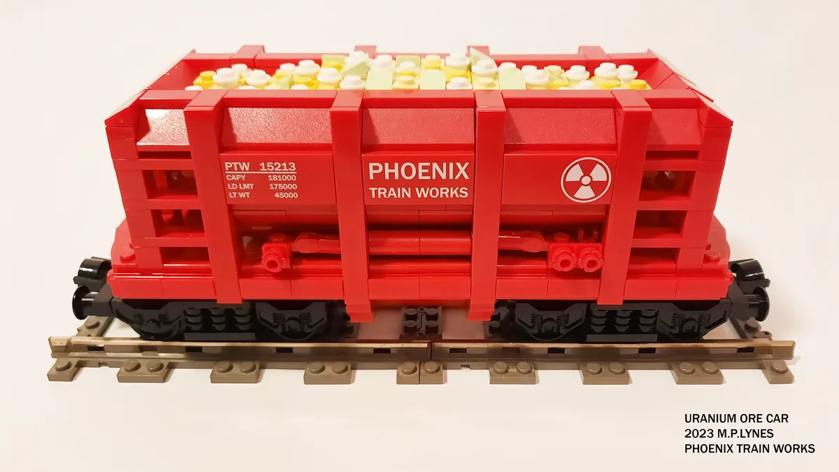 A red LEGO train car labeled 'PHOENIX TRAIN WORKS' with a radiation symbol and capacity markings sits on tracks, filled with…