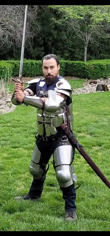 A man with a beard stands in a grassy yard wearing full metallic armor, holding a sword upright in one hand and a long…