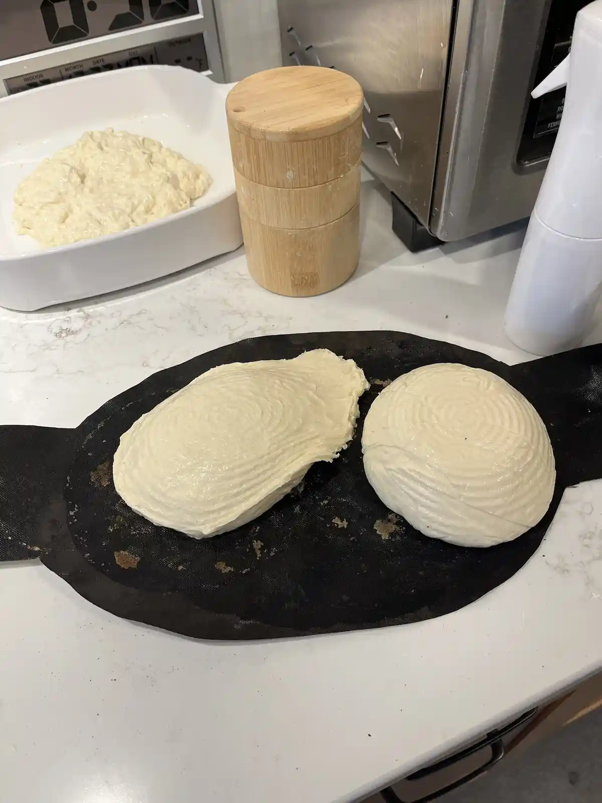 Two pieces of risen bread dough sit on a black silicone mat on a kitchen counter, shaped like a man and a woman. One is a…