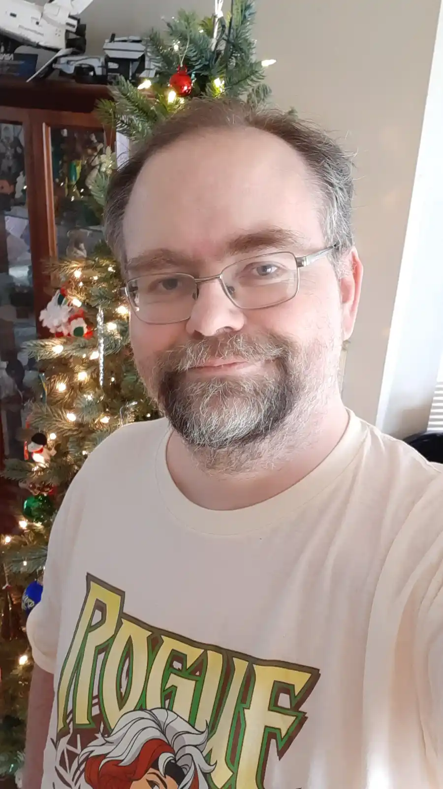 A man with glasses and a graying beard smiles at the camera while standing in front of a lit Christmas tree. He wears a…