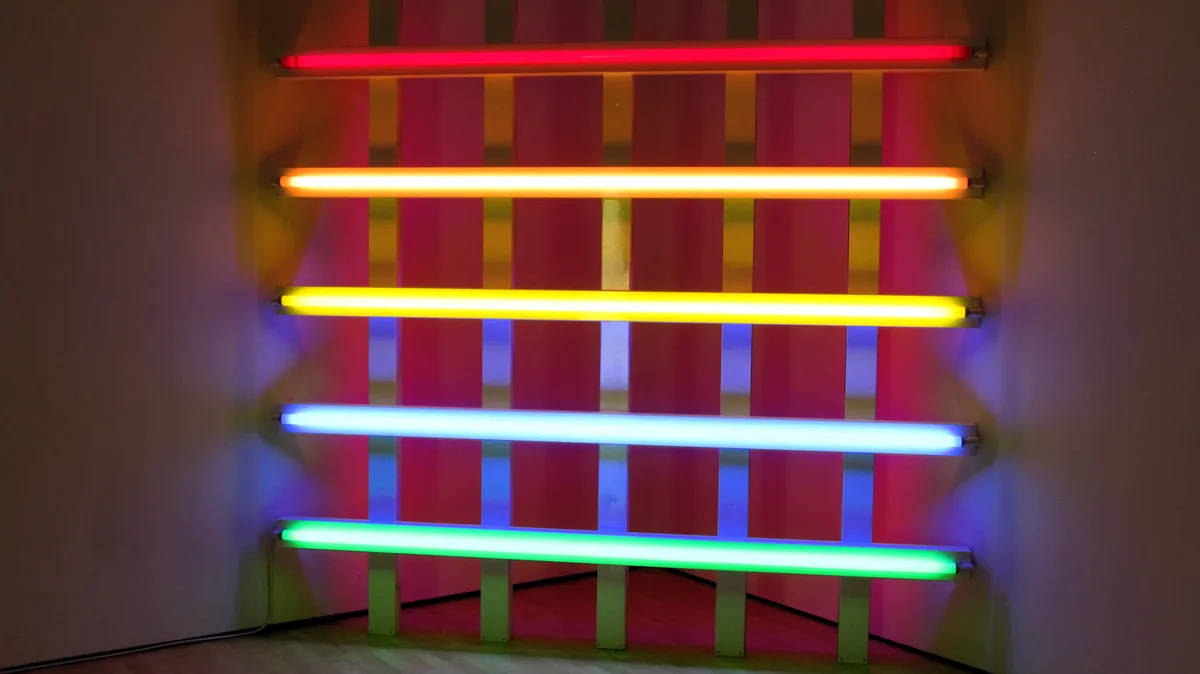 Five horizontal fluorescent light tubes in red, orange, yellow, blue, and green are mounted on a wall with vertical metal…