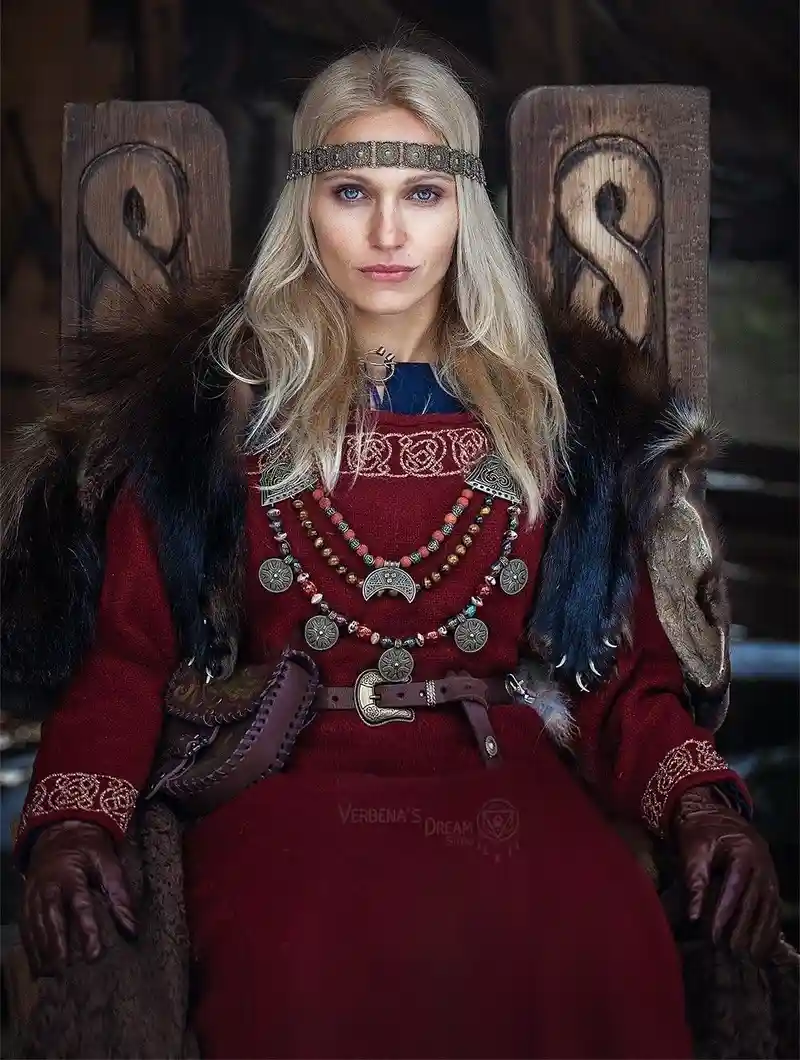 A woman with blonde hair and blue eyes sits regally on a wooden throne adorned with fur, wearing a red embroidered dress…