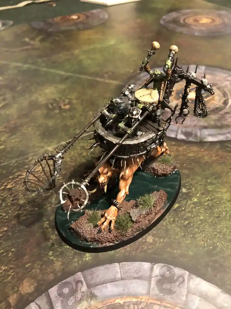 A painted miniature of a fantasy warband riding a monstrous, spiked, mechanical beast on a game board. The overlaid text…