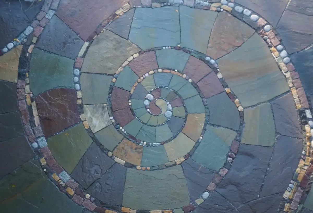 An overhead view of a spiral stone mosaic path, with concentric rings of colored stones leading inward to a central yin-yang…
