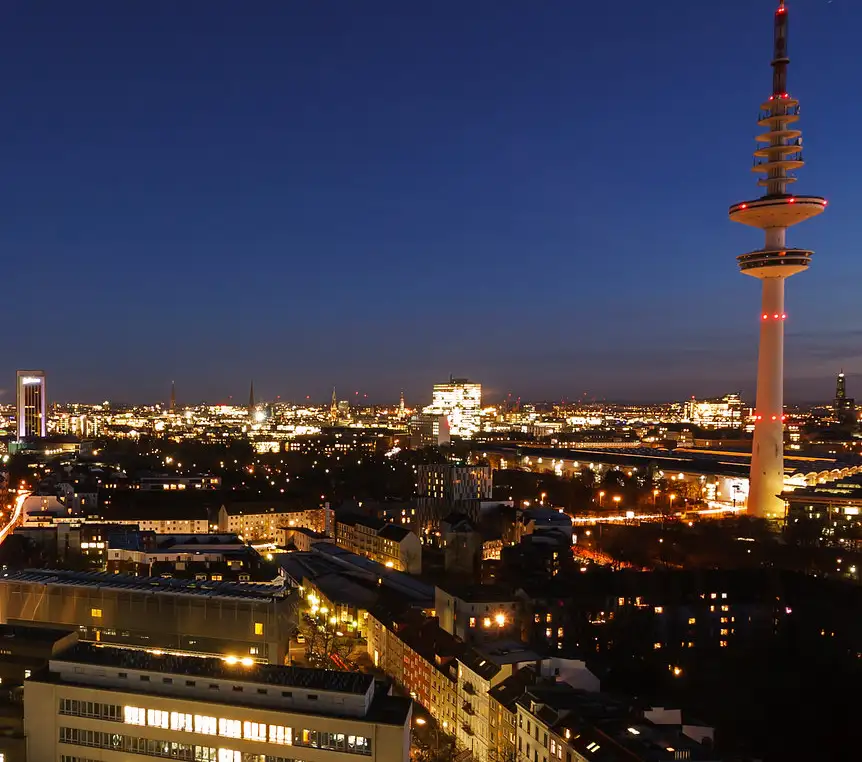 A nighttime cityscape featuring a prominent tall tower with red lights, surrounded by illuminated buildings and streets…