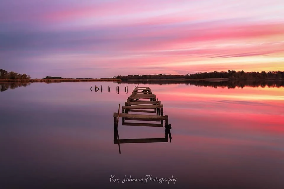 A serene photograph of a weathered wooden pier extending into calm water under a vibrant pink and purple sunset sky, with…