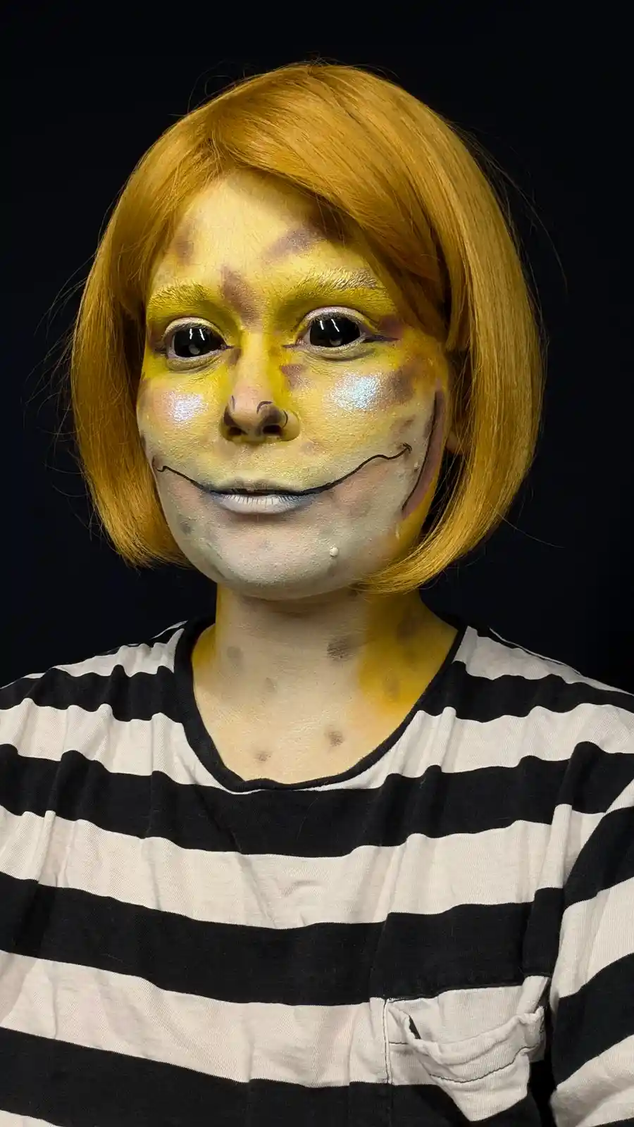 A person with short orange hair and a black-and-white striped shirt has their face painted yellow with brown spots…