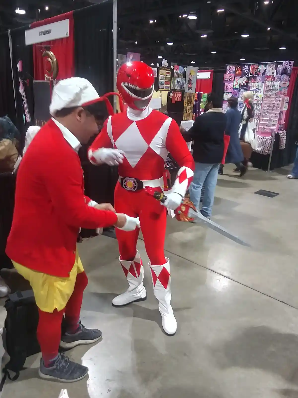 At a convention, a person in a red and yellow costume with a white hat is nervously interacting with a person in a full red…