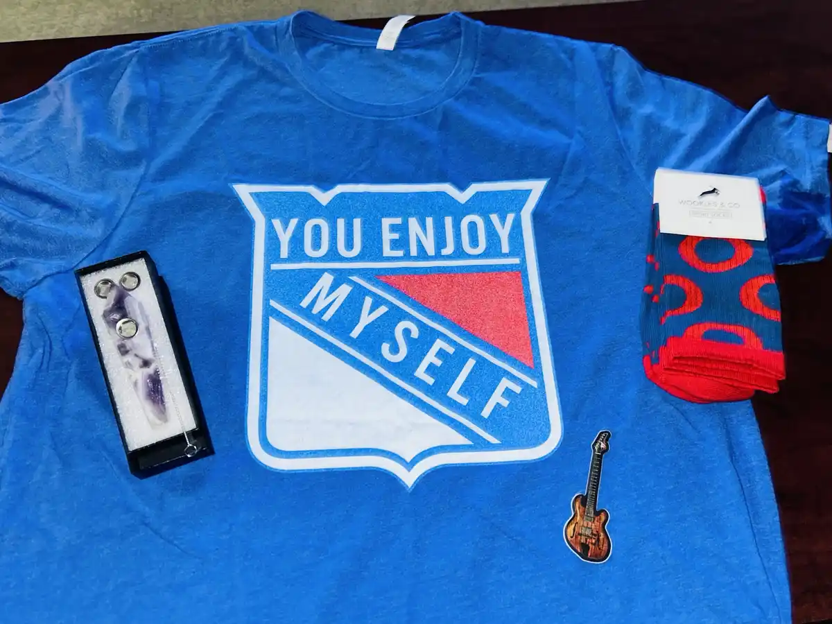A blue t-shirt with a parody NHL logo that reads 'YOU ENJOY MYSELF' is laid out with a packaged crystal, a pair of red and…