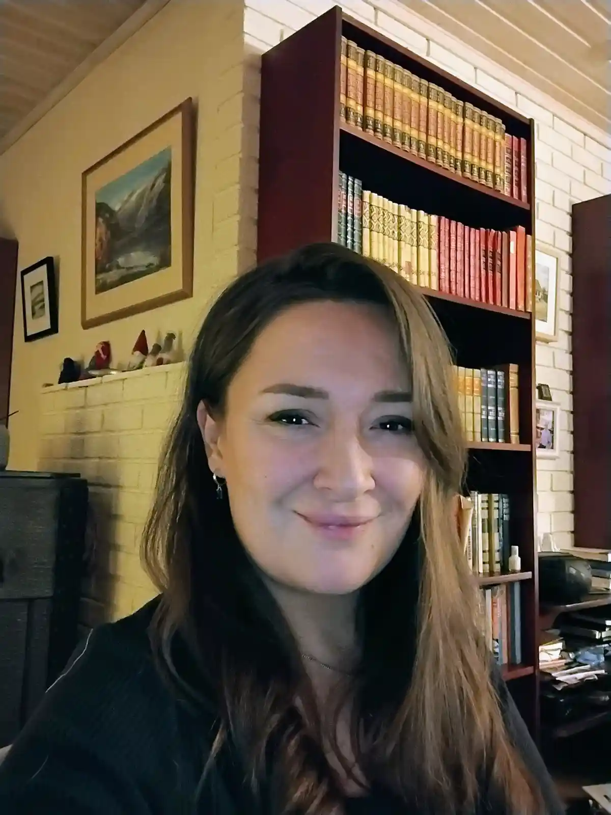 A woman with long brown hair smiles at the camera in a room with a large bookshelf filled with colorful books behind her…